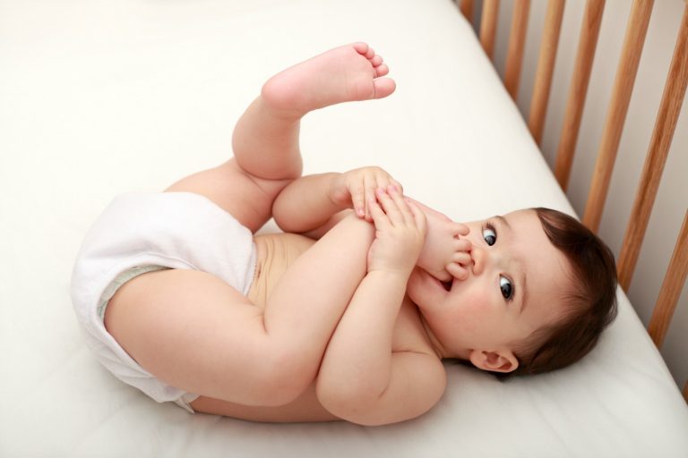 Baby lies on its back in a cot biting one foot and looking at the camera
