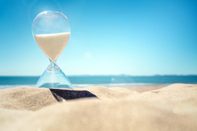 An hourglass on a beach with sand pouring into the bottom against a blue sky