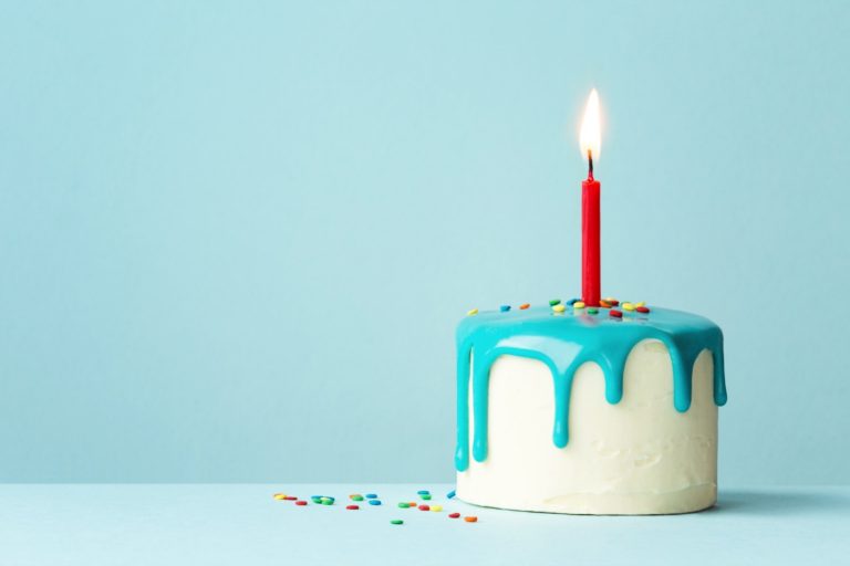White birthday cake with turquoise drip frosting and one red candle against a pale blue background - one year of travelling with a baby