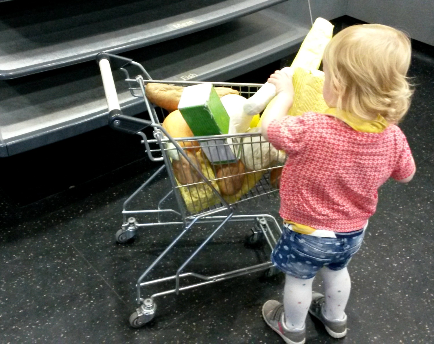 My toddler loading up a mini shopping basket with toy food - a fun day out at Eureka for toddlers