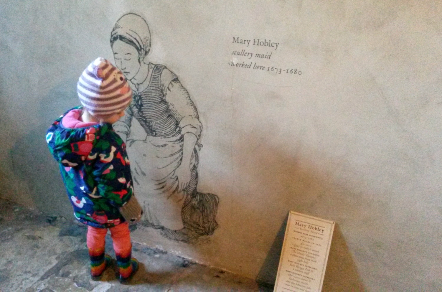 My toddler looks at a picture on the wall below stairs at Ham House in Surrey to show the life and work of one of the maids