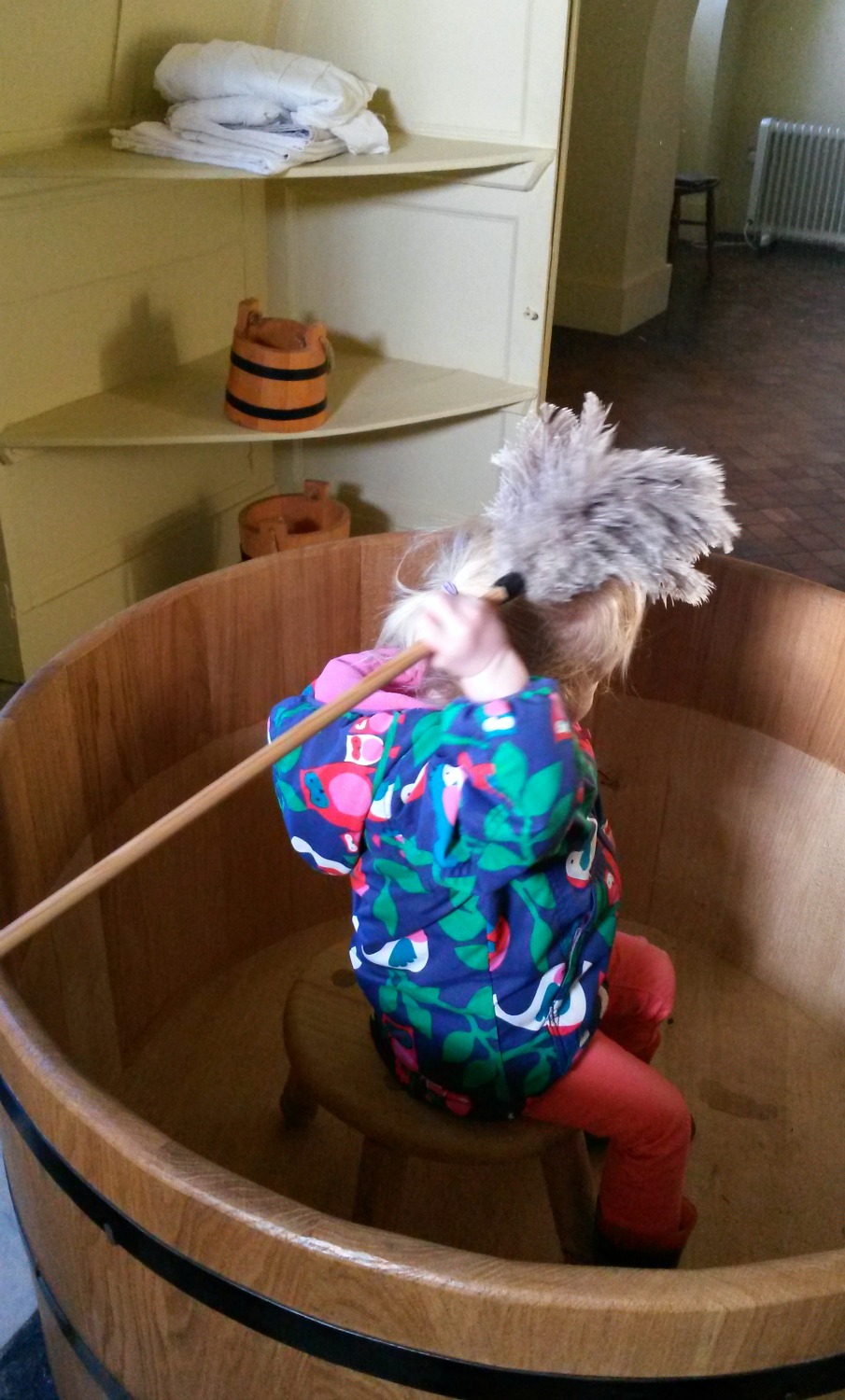 My toddler sitting in an old-fashioned wooden tub pretending to wash herself with a feather duster in the bathroom at Ham House in Surrey