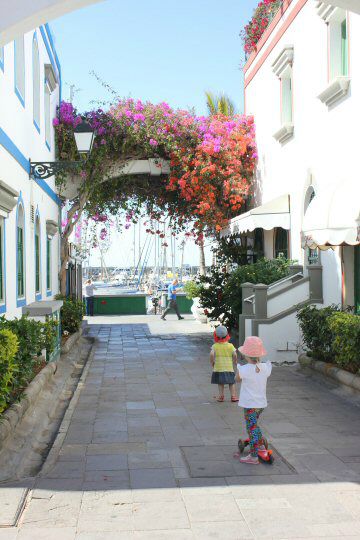 My daughter and her friend with scooter in the streets of Puerto de Mogan, Gran Canaria looking towards a flowery arch above the road