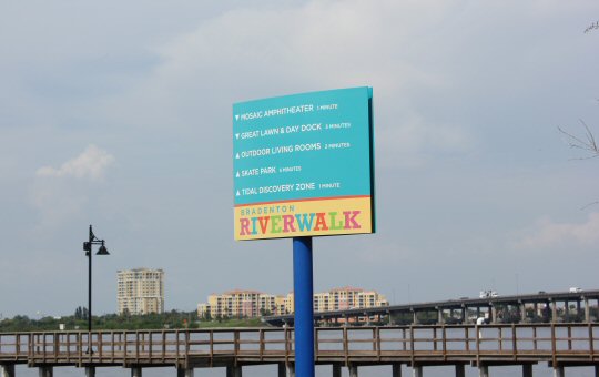 Bright turquoise sign on the Bradenton Riverwalk with details of distances to various local sights and landmarks