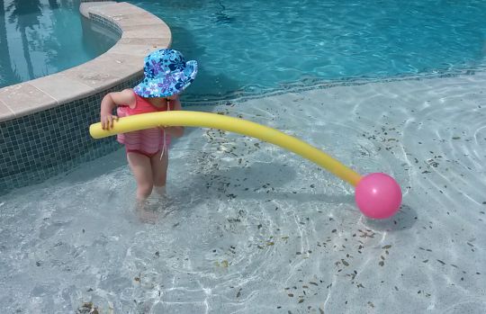 My daughter playing with noodle and ball in the swimming pool while testing her Splashabout floatsuit: review