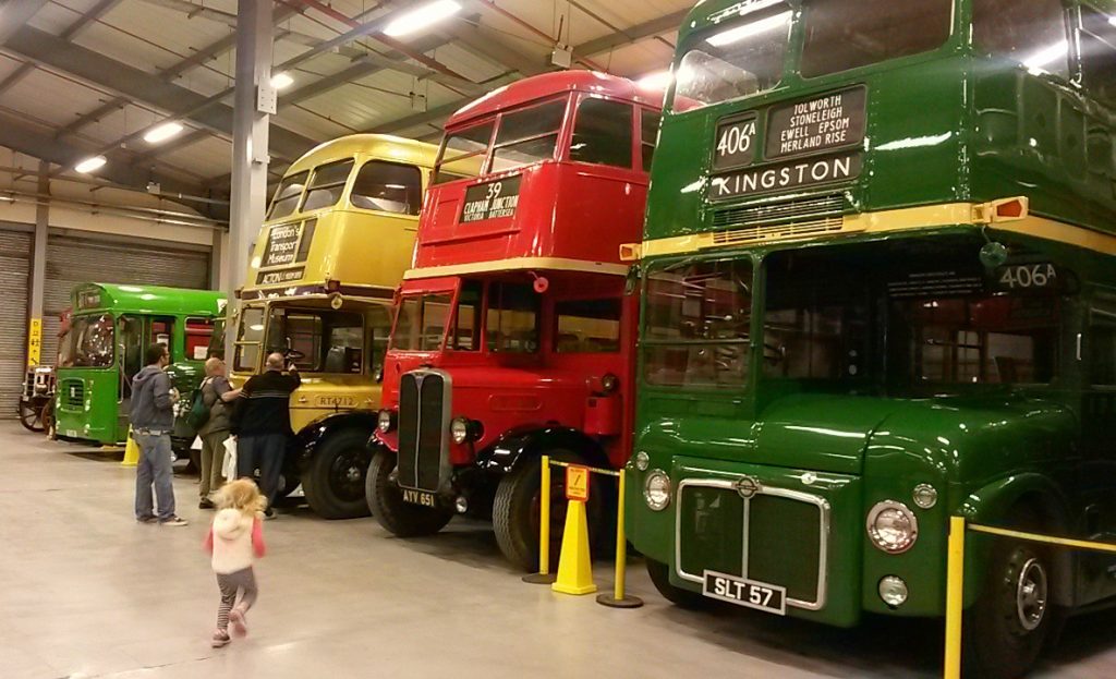 Girl runs towards three vintage buses in yellow, red and green at the London Transport Museum Acton depot