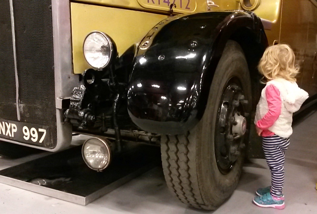 Three year old girl stands next to a huge bus wheel - exploring the London Transport Museum depot with kids