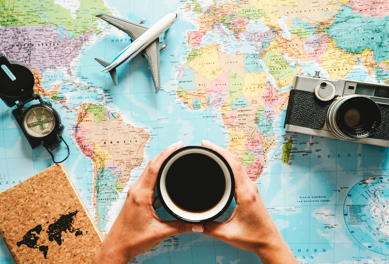 Woman's hands holding coffee on a world map with compass, plane, camera and notebook - my travel resolutions for 2018