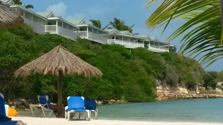 View across the beach with loungers and palapa and some of the accommodation on the cliff above at Verandah Resort: Antigua all inclusive resort