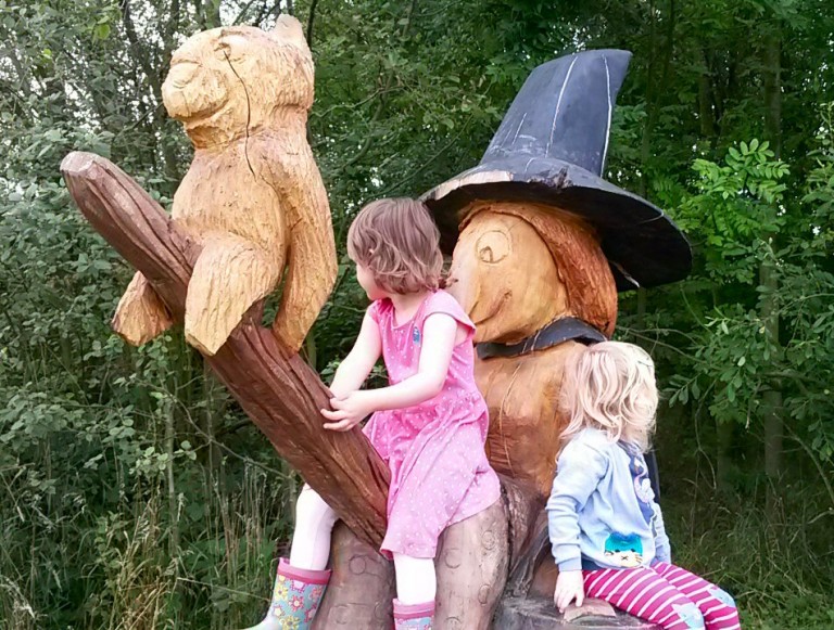 Two girls sit on a wooden broomstick next to a carved statue of a witch from Julia Donaldson's book, on the Room on the Broom trail in Yorkshire