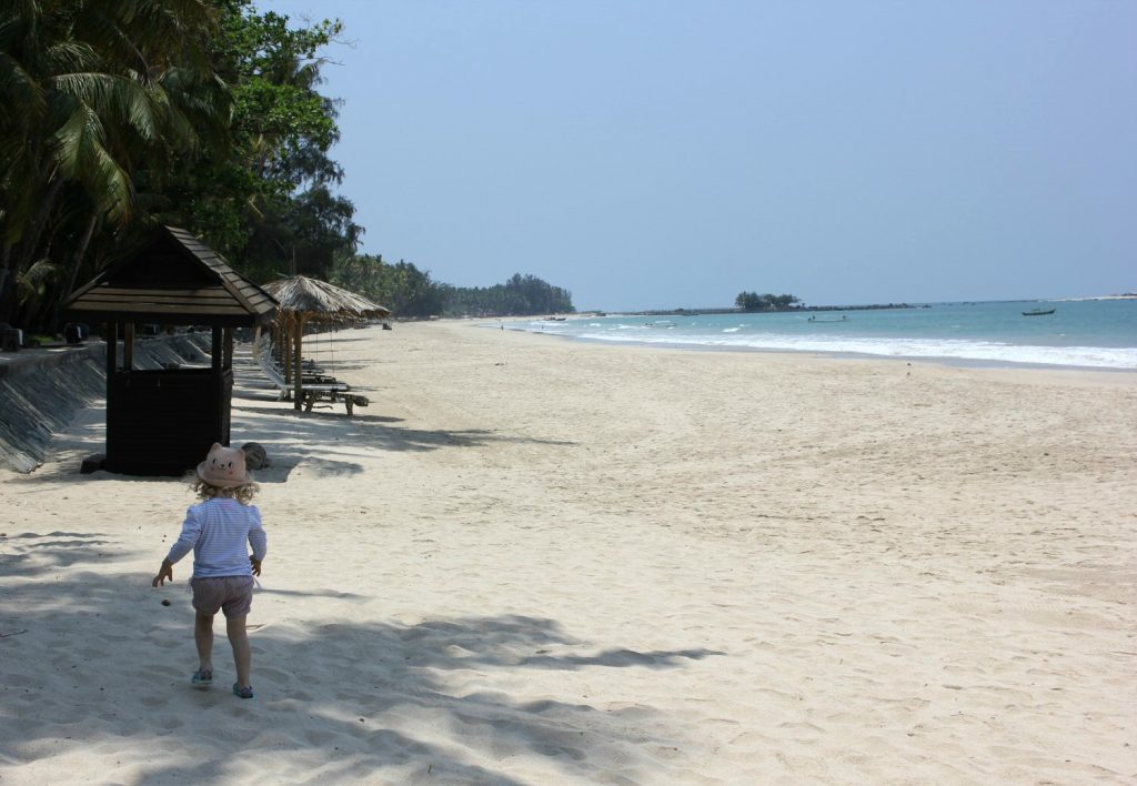 My daughter walks on the sand with trees and palm umbrellas ahead - our Burma beach holiday at Ngapali beach