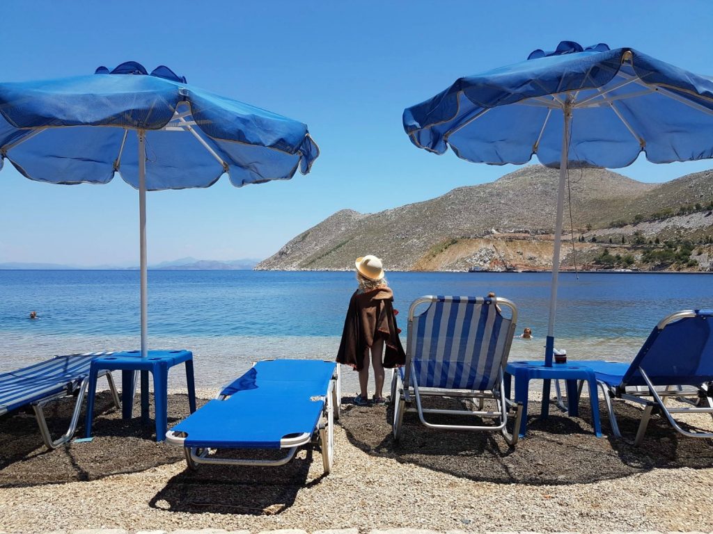 Girl standing by blue loungers and umbrellas on a small beach on Symi in Greece - one of my 13 reasons to go Greek island hopping with kids