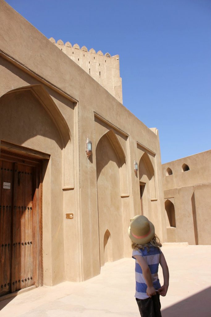My daughter looks up an arched doorways of the sand coloured buildings in Jarbin Castle in Oman against a blue sky- my nine reasons to visit Oman with kids