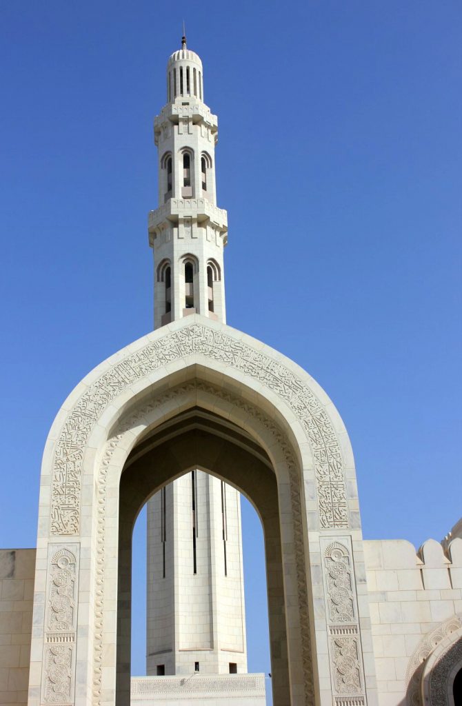 One of the minarets at the Sultan Qaboos Grand Mosque behind a white arch covered with calligraphy in front of a blue sky - my tips for spending 24 hours in Muscat, Oman with kids