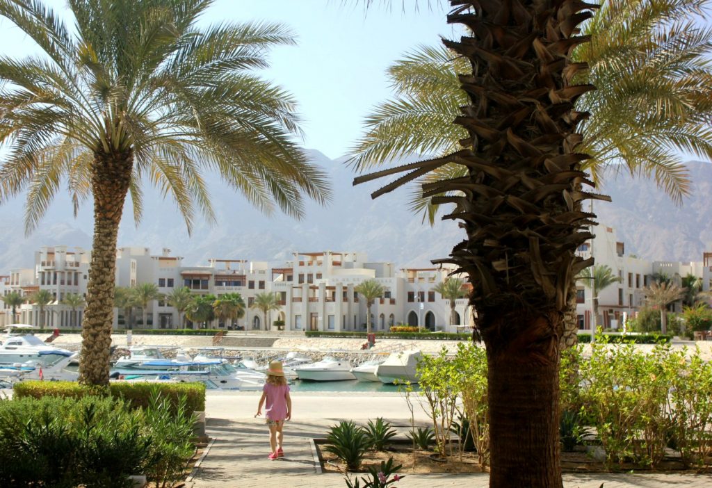 My daughter walks between palm trees at the marina of As Sifa with the white buildings and mountains in the background, not far from Muscat, Oman - my nine reasons to visit Oman with kids