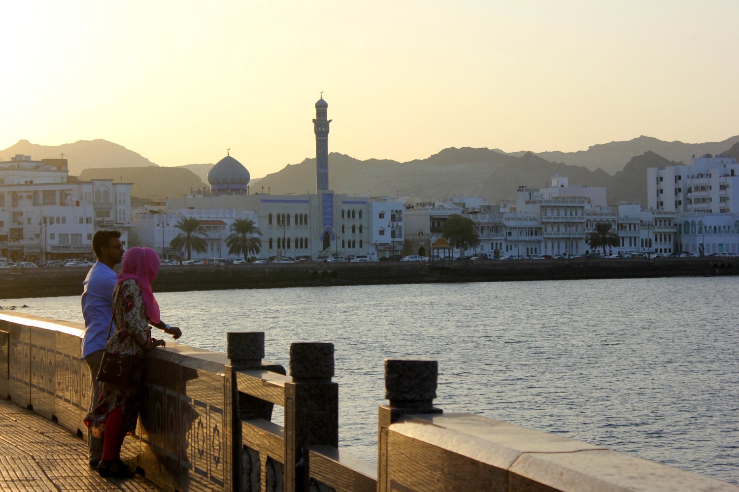 A view from the Corniche at Mutrah towards the sea and Muscat, a couple silhoutted against the sunset - how to explore the sprawling capital was one of my Oman travel lessons learned