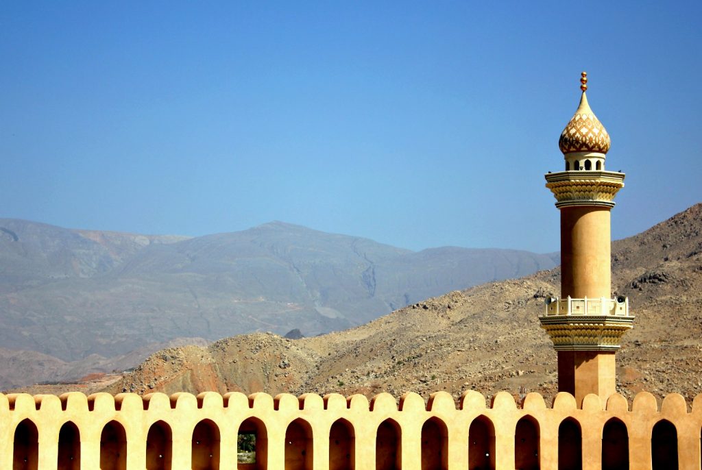 A view from Nizwa fort out onto the former capital of the interior in Oman, looking out across the golden arched fortifications to the minaret tower and Hajar mountains beyond - my nine reasons to visit Oman with kids