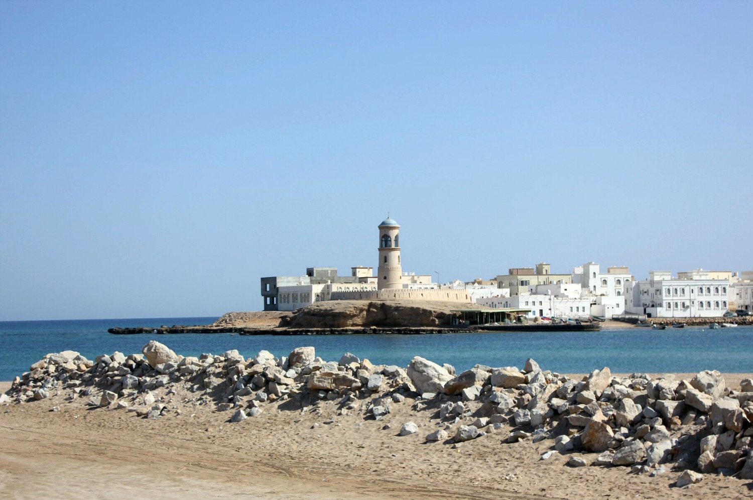 Looking across the blue water to the white and sand coloured lighthouse and buildings of Sur in Oman - my nine reasons to visit Oman with kids
