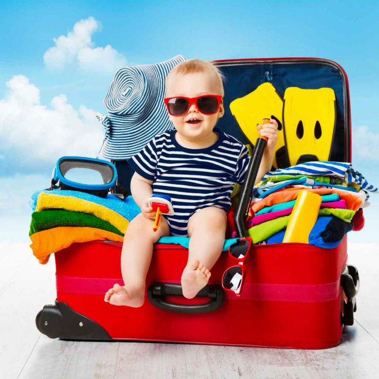 Baby sitting in a suitcase packed with clothes and holiday things, blue sky and clouds behind - my essential baby packing list for travel