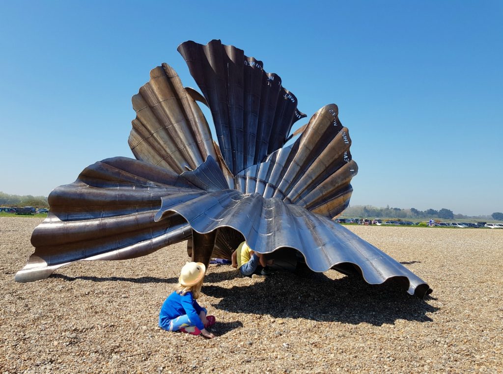 My daughter plays on the shingle in front of Maggi Hambling's scallop sculpture on the beach at Aldeburgh - one of my things to do in Aldeburgh with kids if you're visiting Suffolk