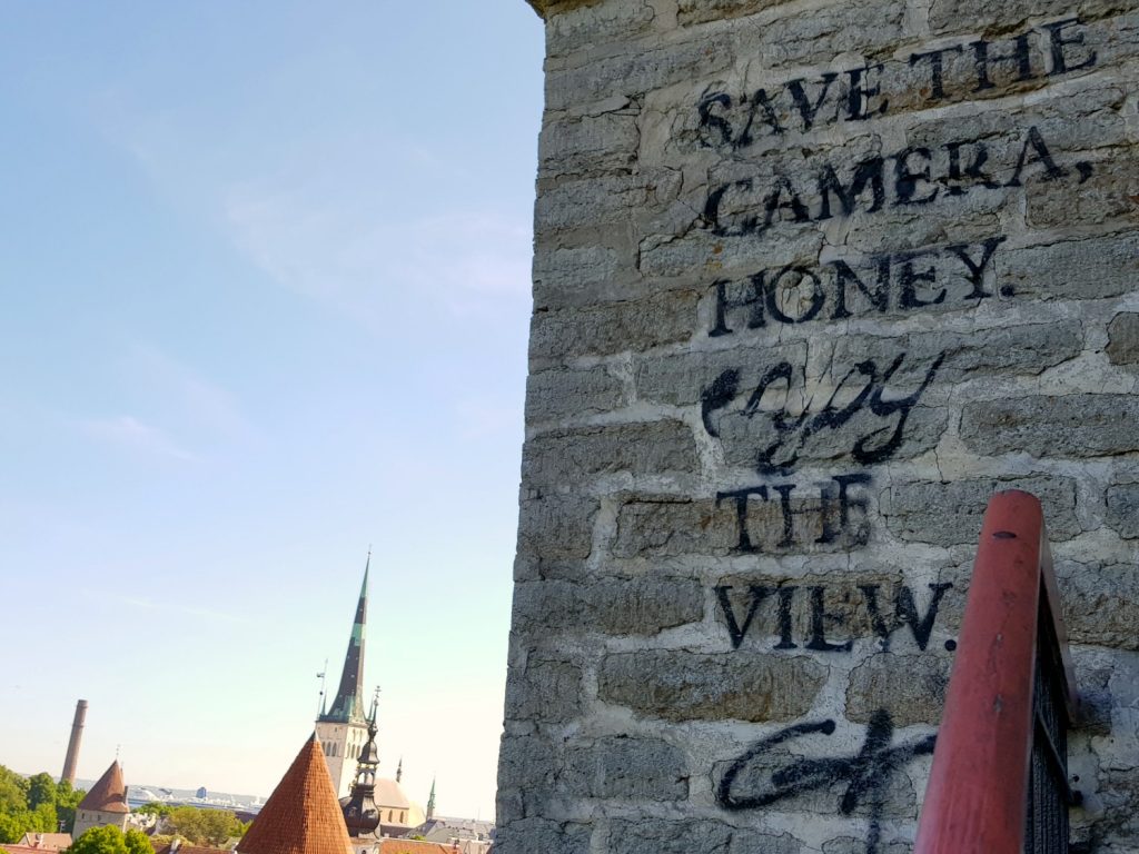 Graffiti reading 'save the camera honey, enjoy the view' with the spires of Tallinn in the background - the city's views are just one of the many reasons to visit Tallinn, Estonia