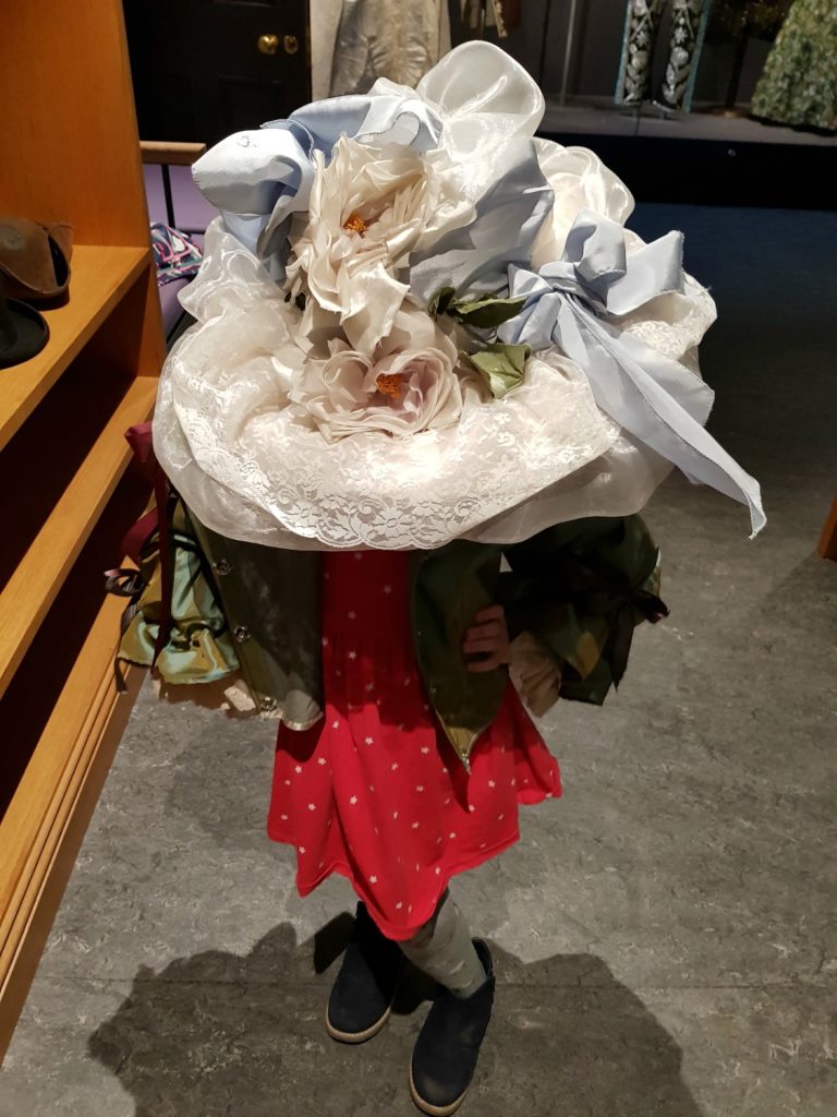 My daughter under a gigantic picture hat, playing dress up in Victorian clothes in the fashion gallery - my review of a free day out in London, visiting Gunnersbury Park Museum with kids