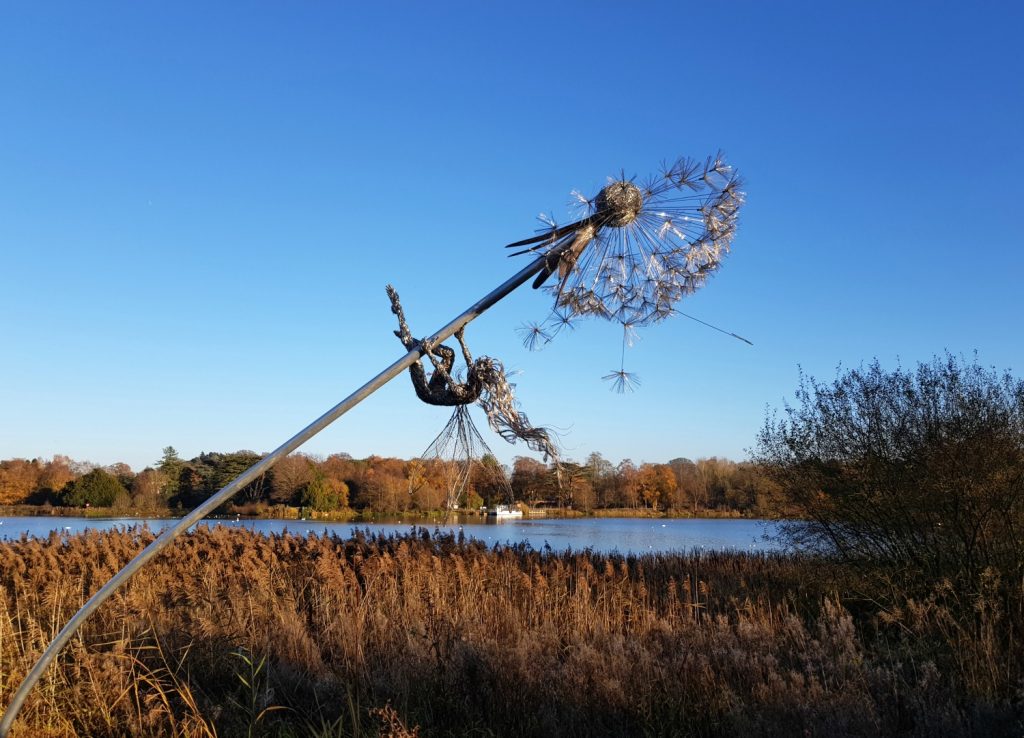 Wire fairy sculpture hanging off a wire dandelion clock against a blue sky with lake in the background - going wild in Stoke-on-Trent and following the Trentham fairy trail