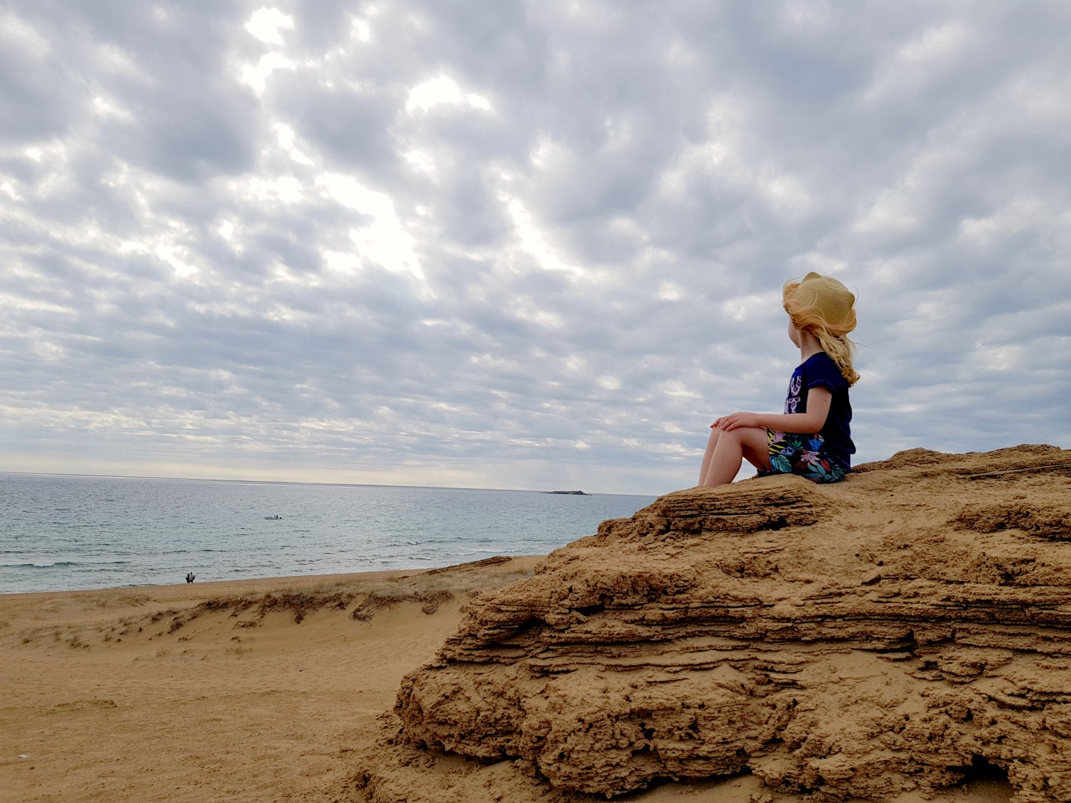 My daughter sits on a rock above the sand and gazes out to the cloudy skies and sea at Issos beach - one day in Corfu exploring some of the island in an Ikos Dassia mini