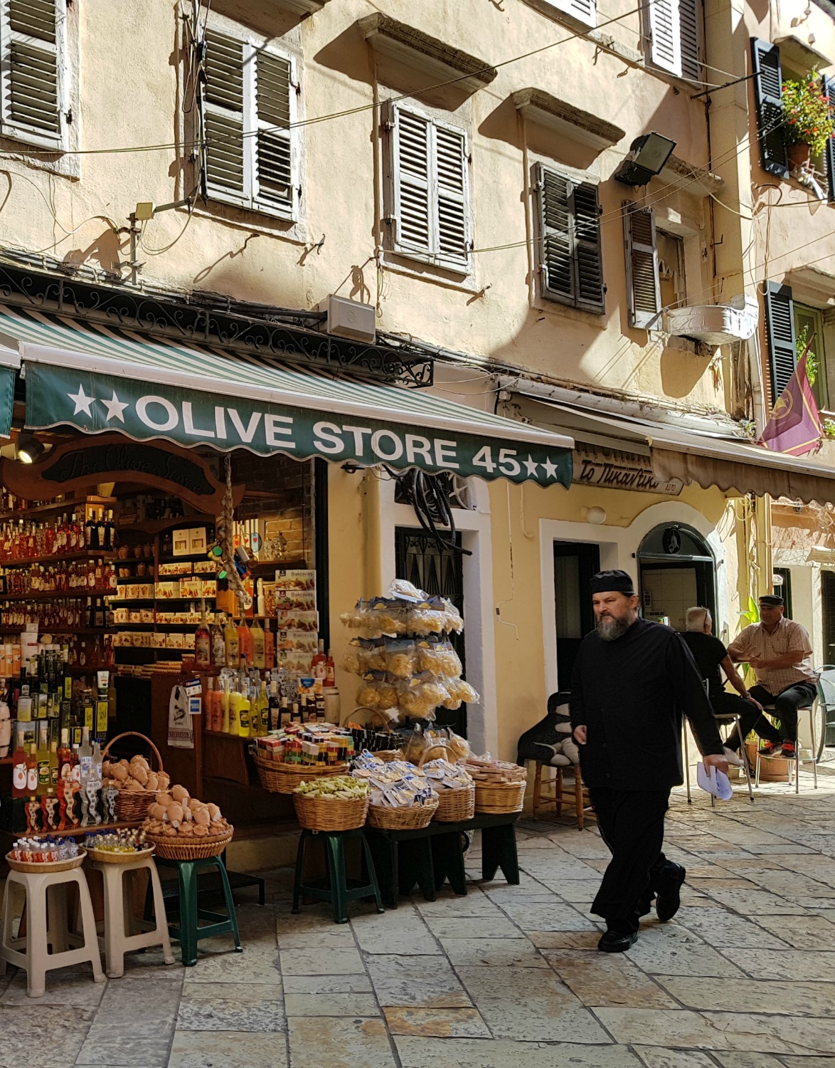 A black clad Greek Orthodox priest walks past an olive shop - three things to do in Corfu Town with kids