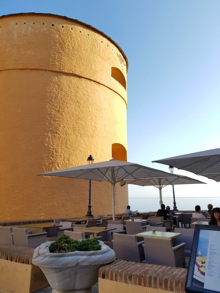 The huge deep yellow round tower of the citadel at Bastia, Corsica, with small cafe tables under umbrellas in front - my Corsica short break visiting Bastia and Calvi in a long weekend