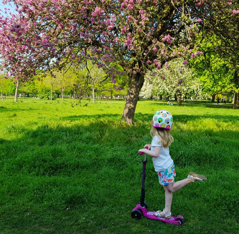 My daughter poses on her Maxi Micro scooter in front of a tree with pink blossom - choosing the best scooter for kids