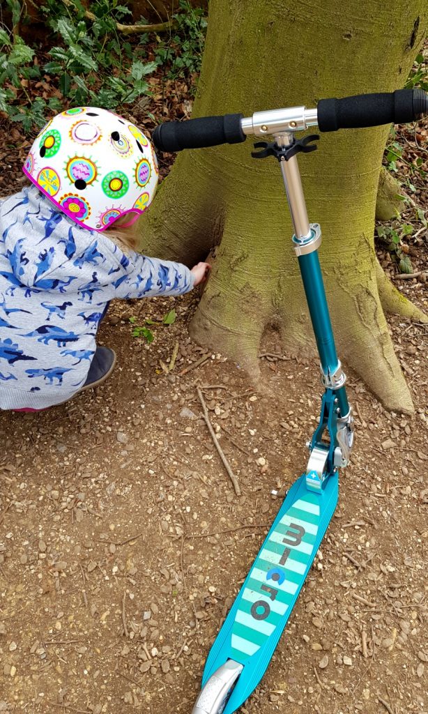 My daughter crouches next to her Micro Sprite scooter looking for a fairy door in a tree on the Bath Skyline Family Discovery trail
