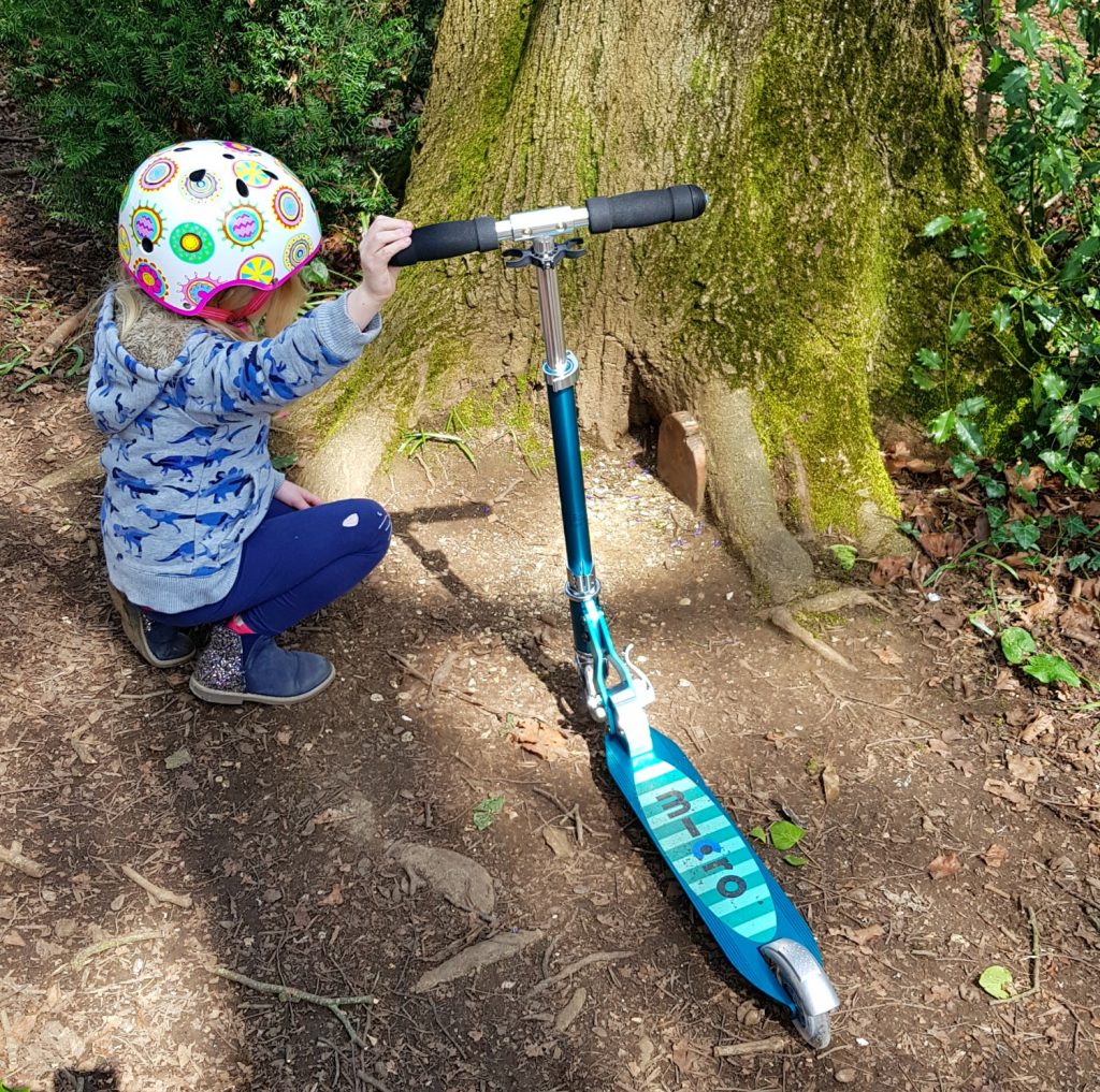My daughter holding her Micro Sprite scooter crouches down next to a fairy door in a tree on the Bath Skyline Family Discovery trail