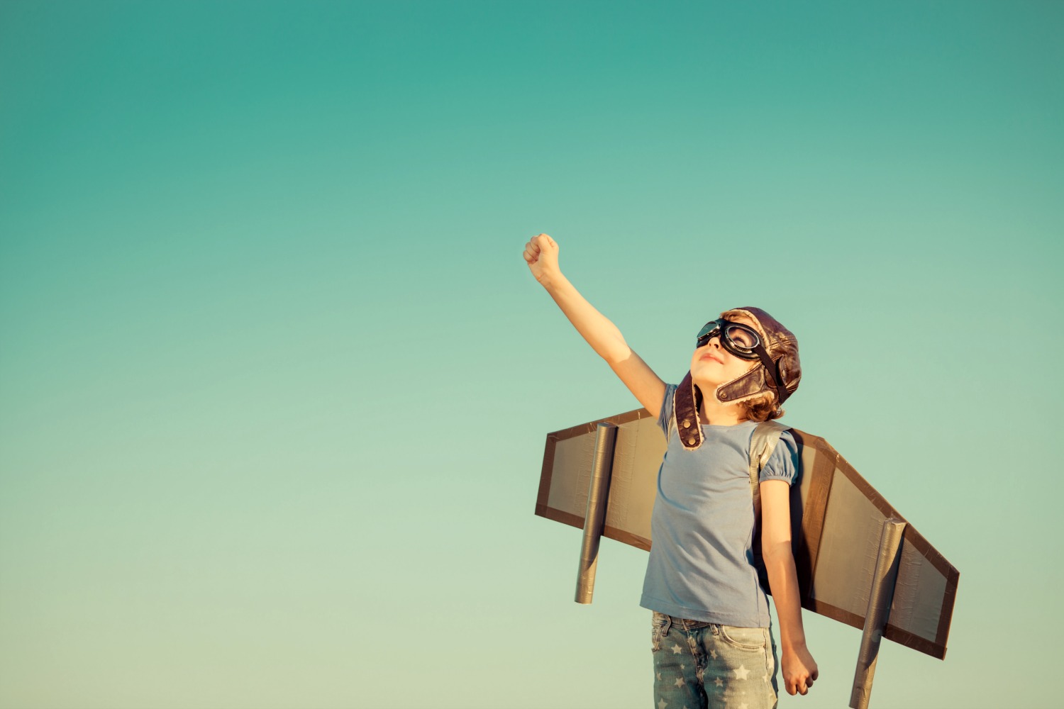 Child with toy wings and old-fashioned pilot goggles reaches up to the sky - launching the Kidventures family travel podcast