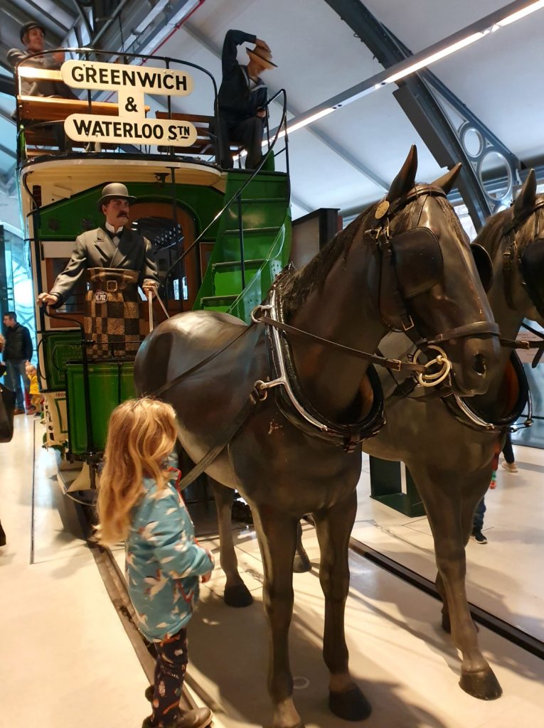 My daughter next to model horses pulling an early tram at the London Transpofrt Museum