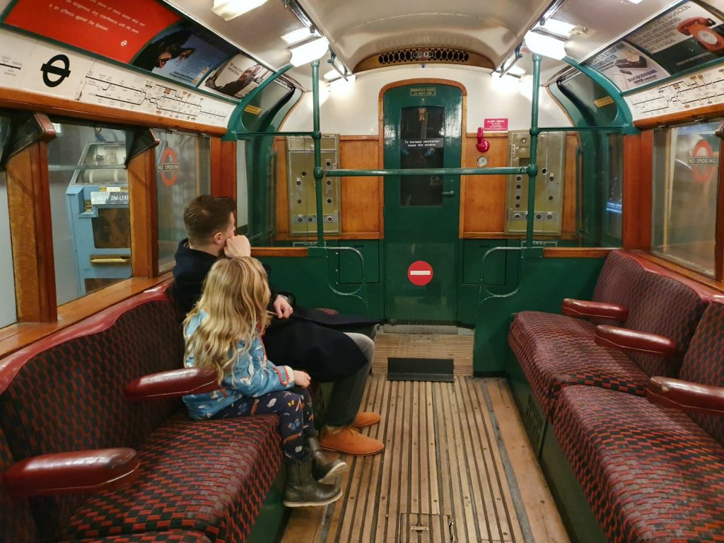 My daughter and husband on board a vintage London Underground train with fraying moquette seats and grooved wooden floor at the London Transport Museum