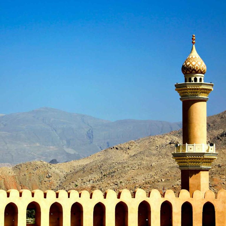 A view from Nizwa fort out onto the former capital of the interior in Oman, looking out across the golden arched fortifications to the minaret tower and Hajar mountains beyond - my nine reasons to visit Oman with kids