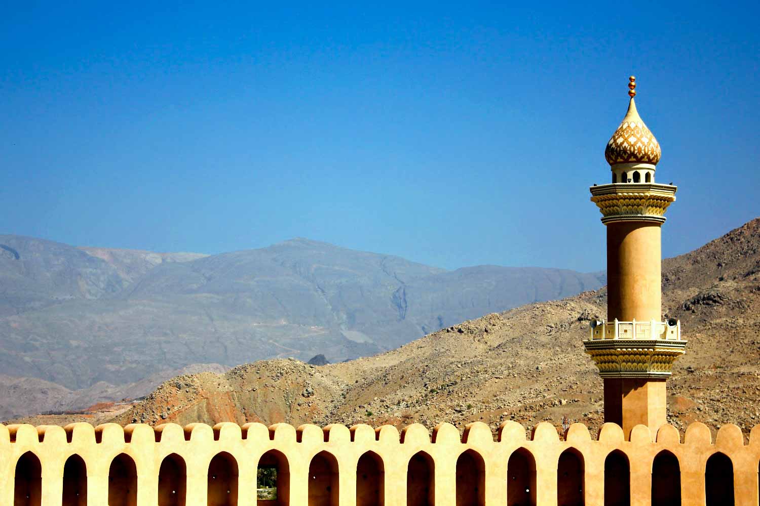 A view from Nizwa fort out onto the former capital of the interior in Oman, looking out across the golden arched fortifications to the minaret tower and Hajar mountains beyond - my nine reasons to visit Oman with kids