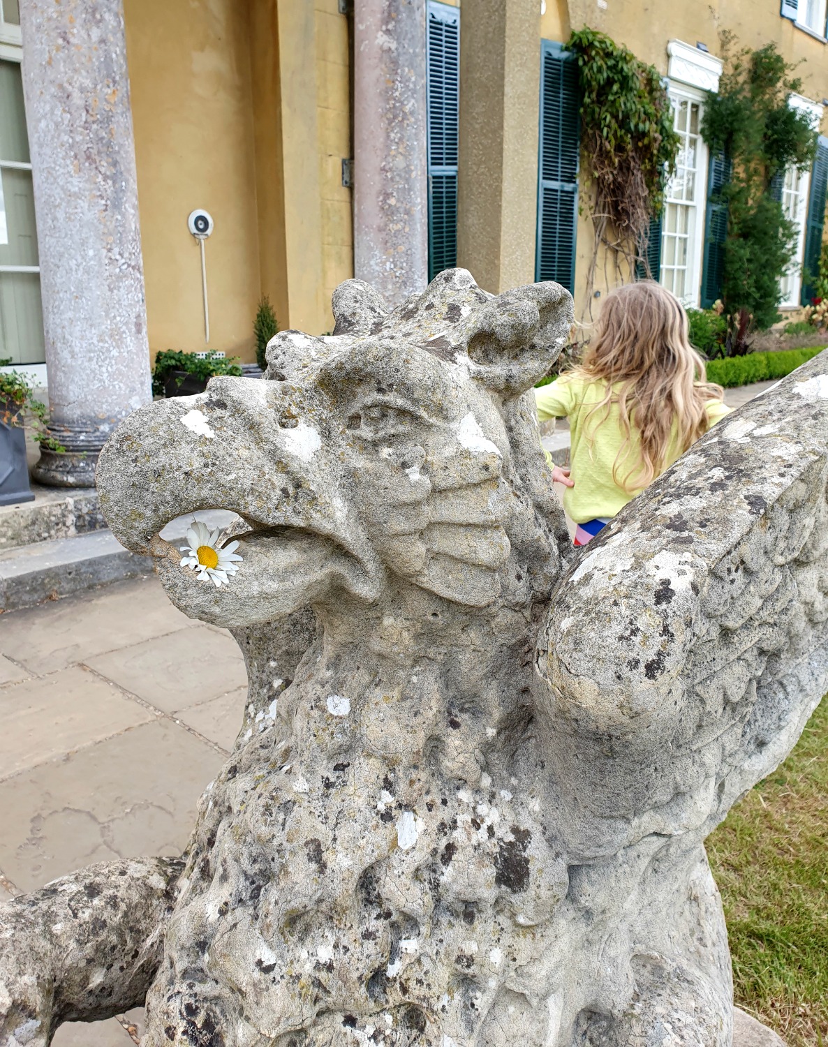 My daughter by a stone statue of a griffin, having fed it a giant daisy during out visit to Polesden Lacey after lockdown