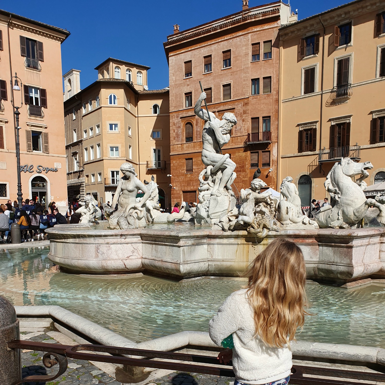 My daughter looks at a fountain in the Piazza Navona, Rome with some of the ochre and golden buildings in the background against a blue sky