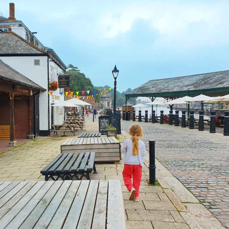 My daughter walks along the cobbled street of the quayside with old buildings and the waterfront in the distance - exploring Exeter with kids