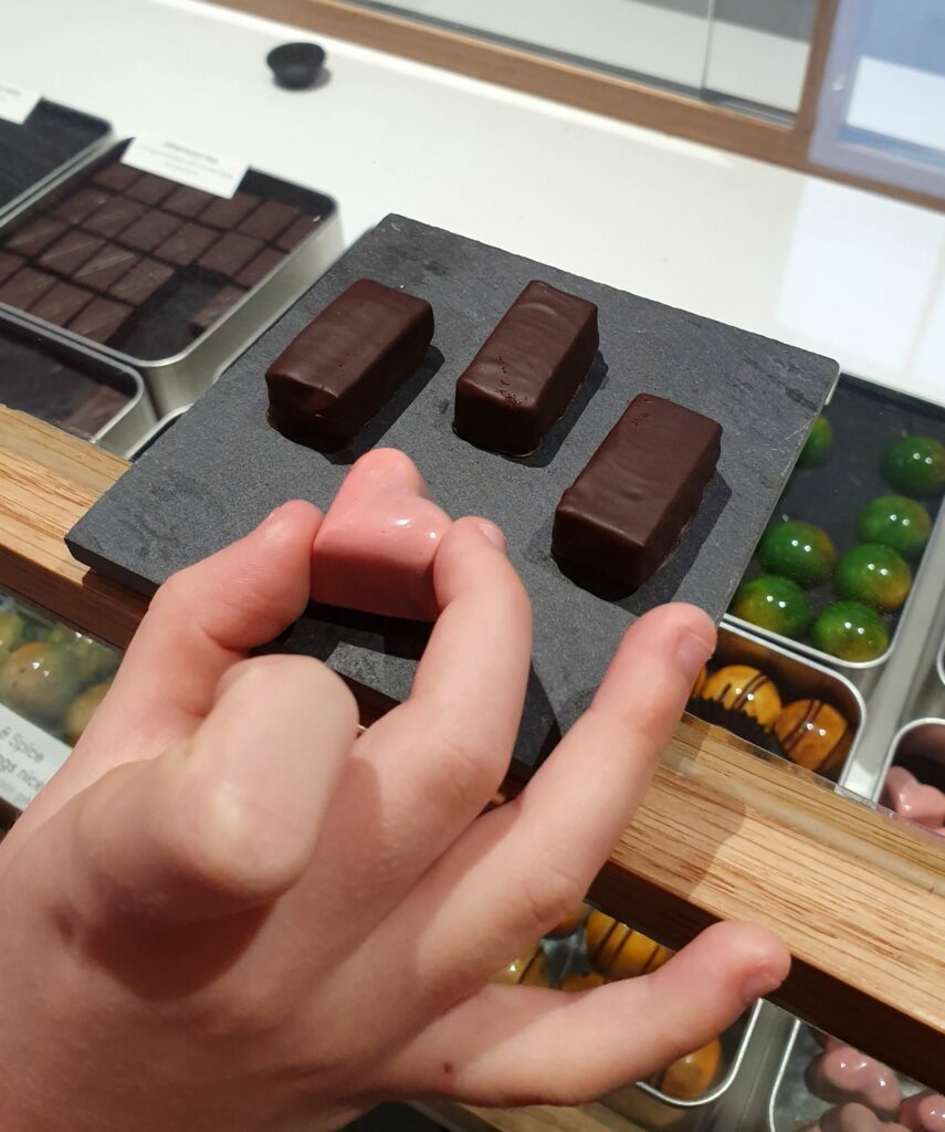 My daughter's hand picks up a raspberry heart-shaped chocolate with other saffron decorated chocolates on a piece of slate in the background, during a visit to Hill Street Chocolate in Saffron Walden