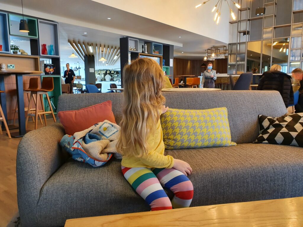 My daughter sits on a sofa with colourful cushions in the lobby at Holiday Inn Winchester - one of the best places to stay in Winchester with kids