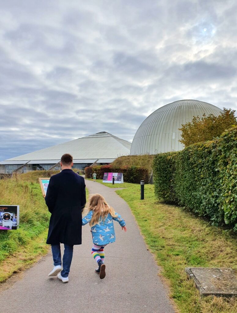 My husband and daughter walk towards Winchester Science Centre with the dome of the Planetarium visible under a cloudy sky - a great day out in Winchester in the rain