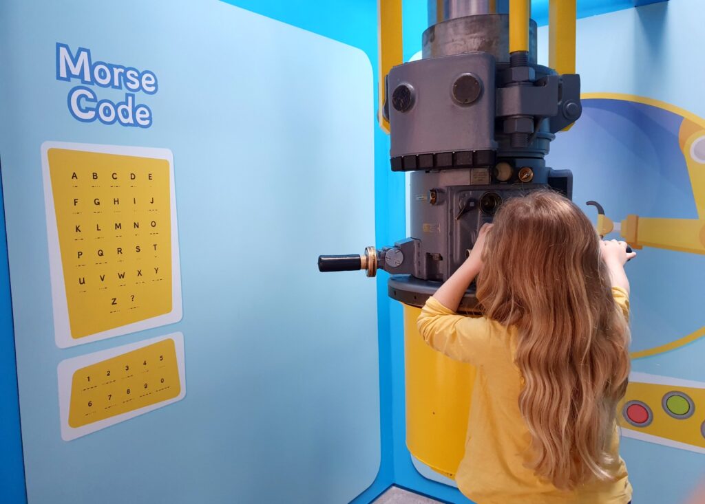 My daughter looks through a periscope next to a Morse Code chart in the whale-themed section of Winchester Science Centre
