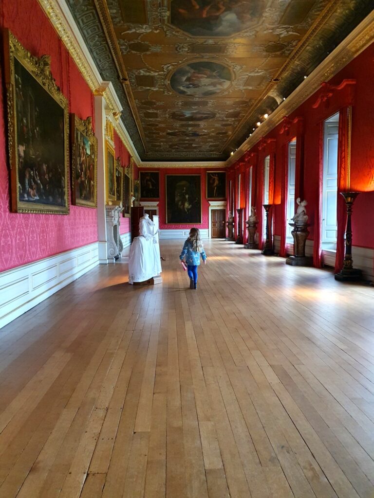 View of the length of the King's Gallery with its red walls, painted ceiling and artwork, as my daughter looks at a recreated dress on display, during a visit to Kensington Palace with kids
