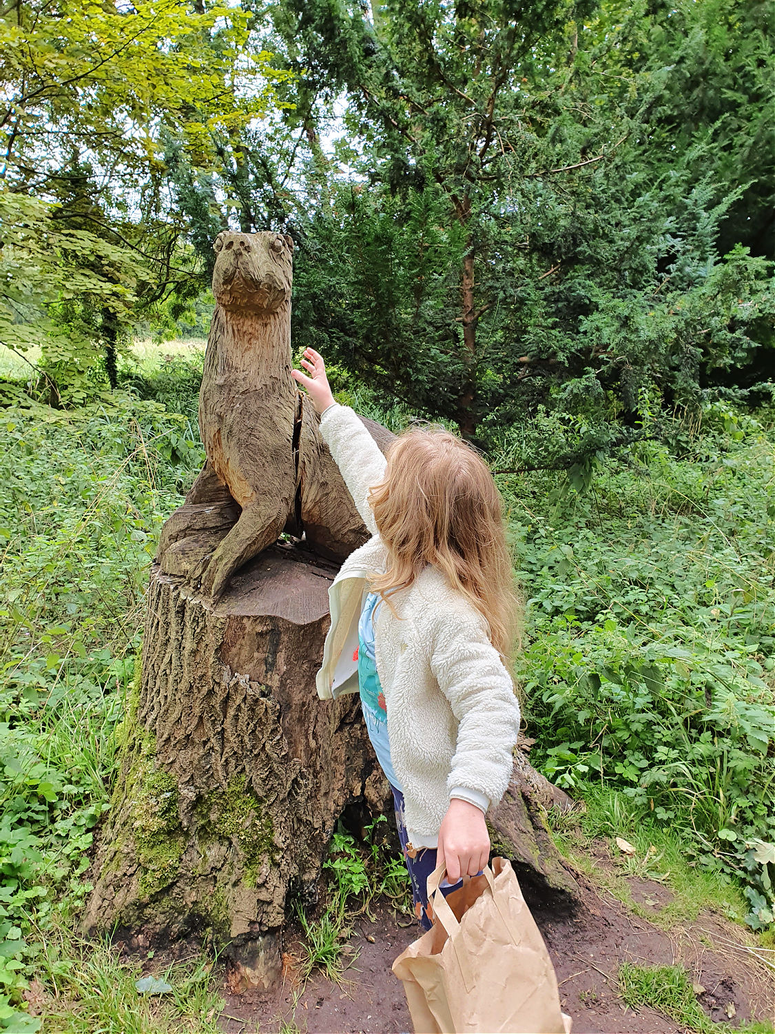 My daughter reaches to a wooden otter sculpture in the woods at Oxburgh Hall in Norfolk - a day out at Oxburgh Hall with kids