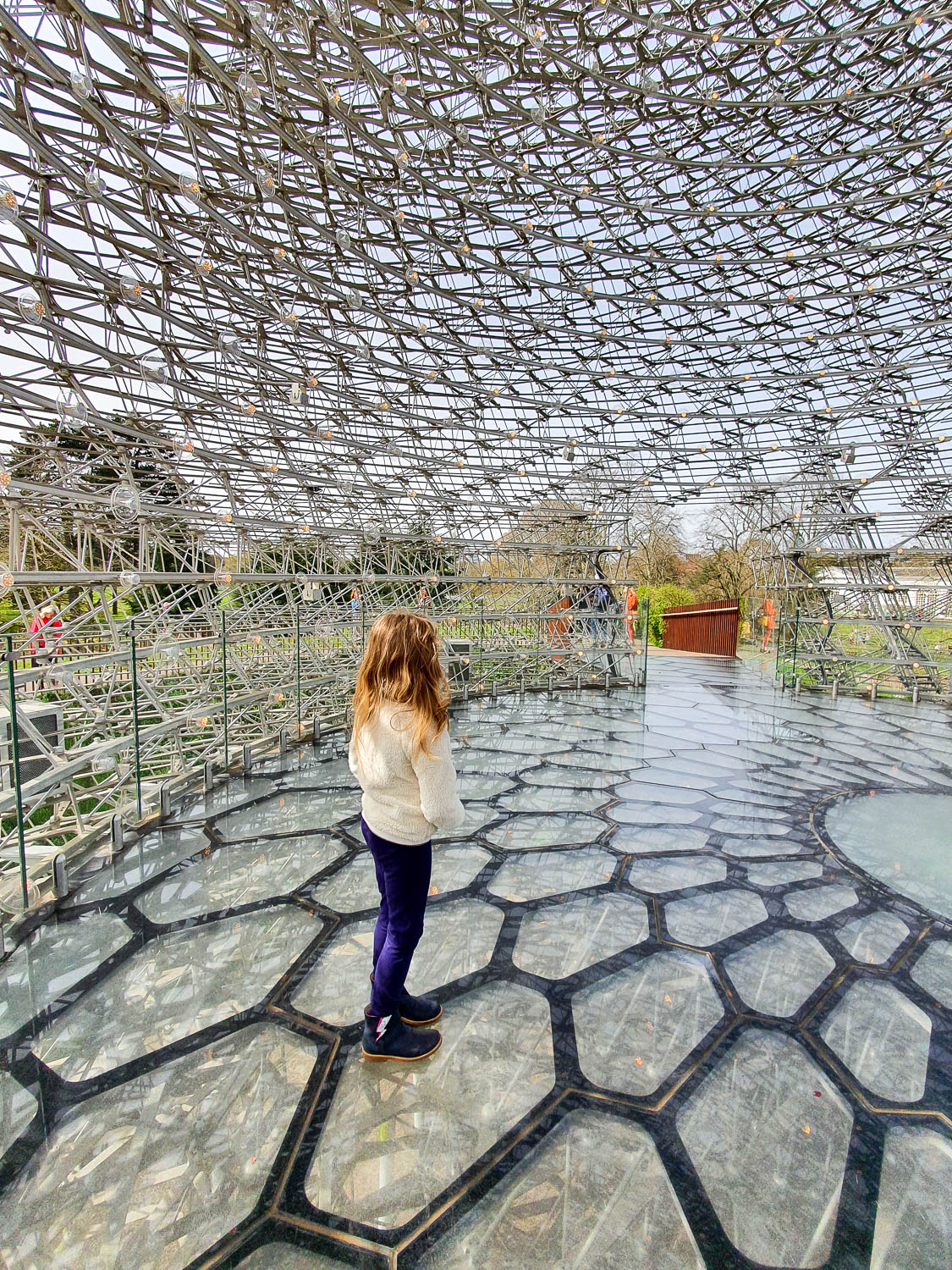 My daughter stands inside The Hive sculpture at Kew Gardens with hexagon shapes on the floor and the metal structure above - a family day out at Kew Gardens in spring