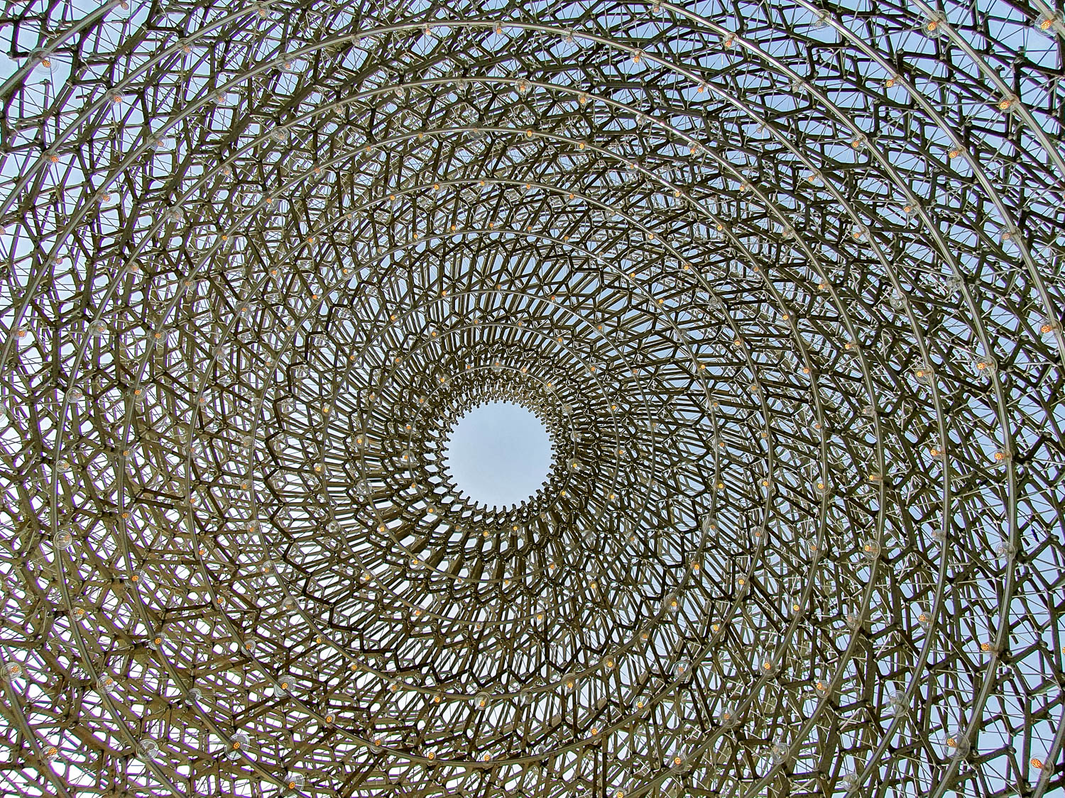 View of the swirling spiralling metal structure of the Hive at Kew Gardens with its circular opening in the roof showing blue sky - a family day out at Kew Gardens