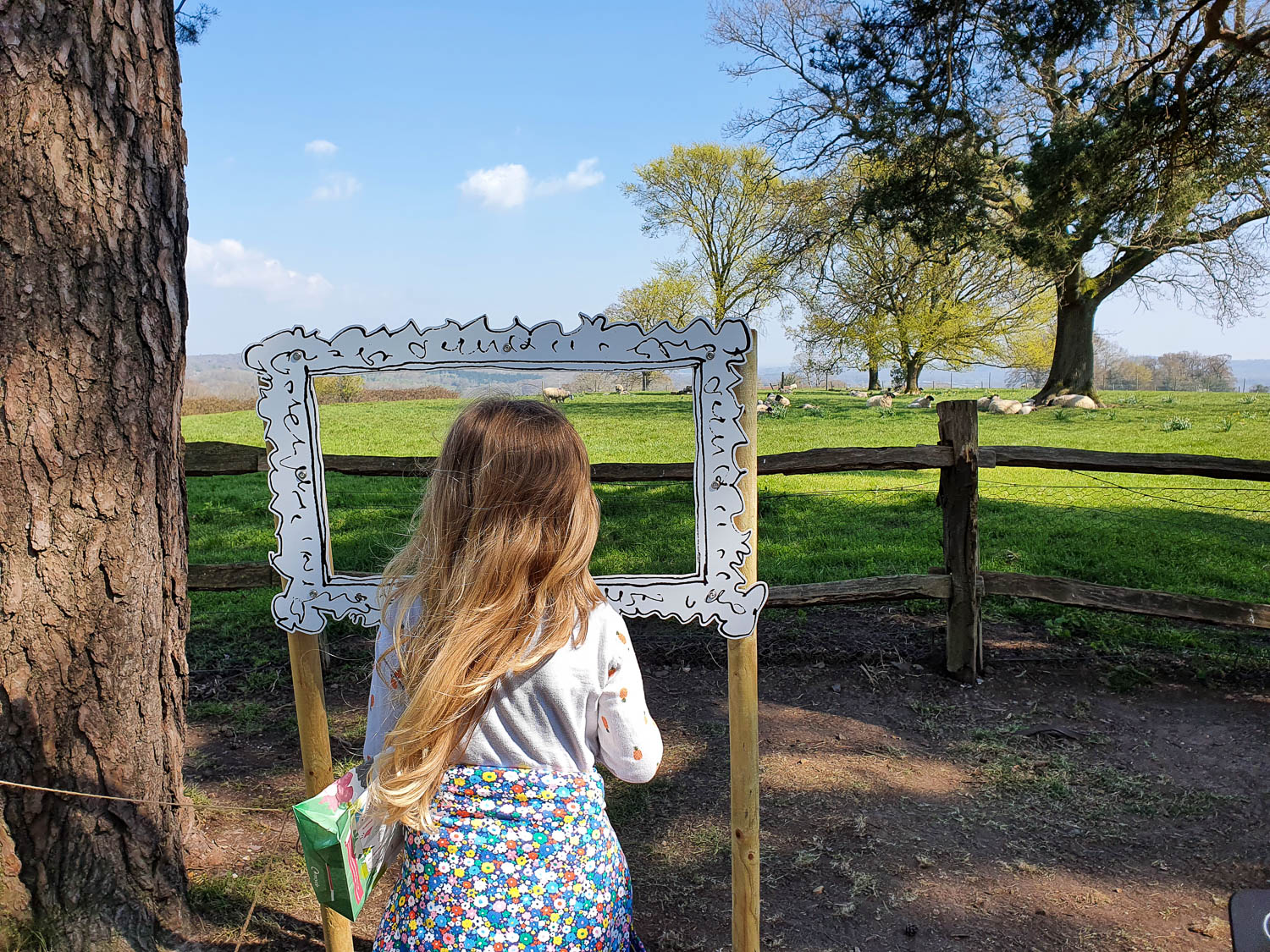 My daughter looks through a frame towards a field of sheep and a distance viaduct as part of a National Trust family trail for Easter - a day out at Nymans with kids